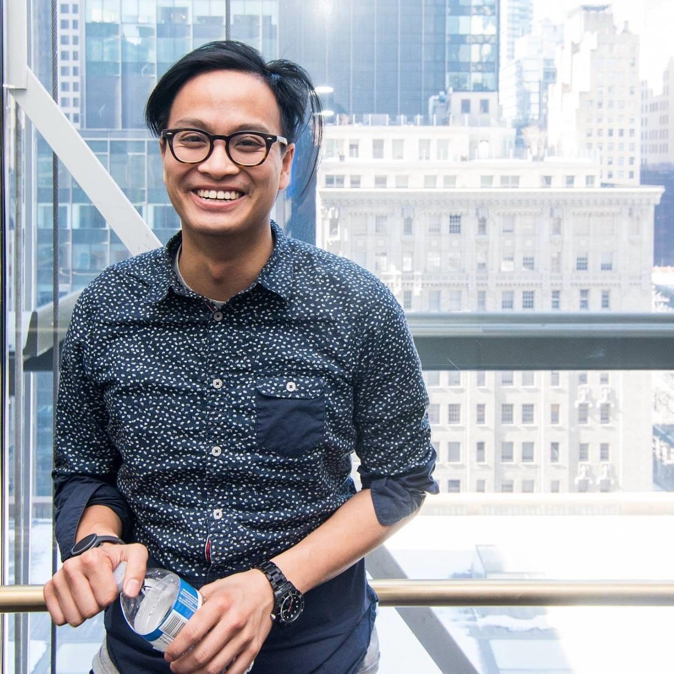 Phi Pham smiling in a dotted button-down shirt, in front of a window over Manhattan